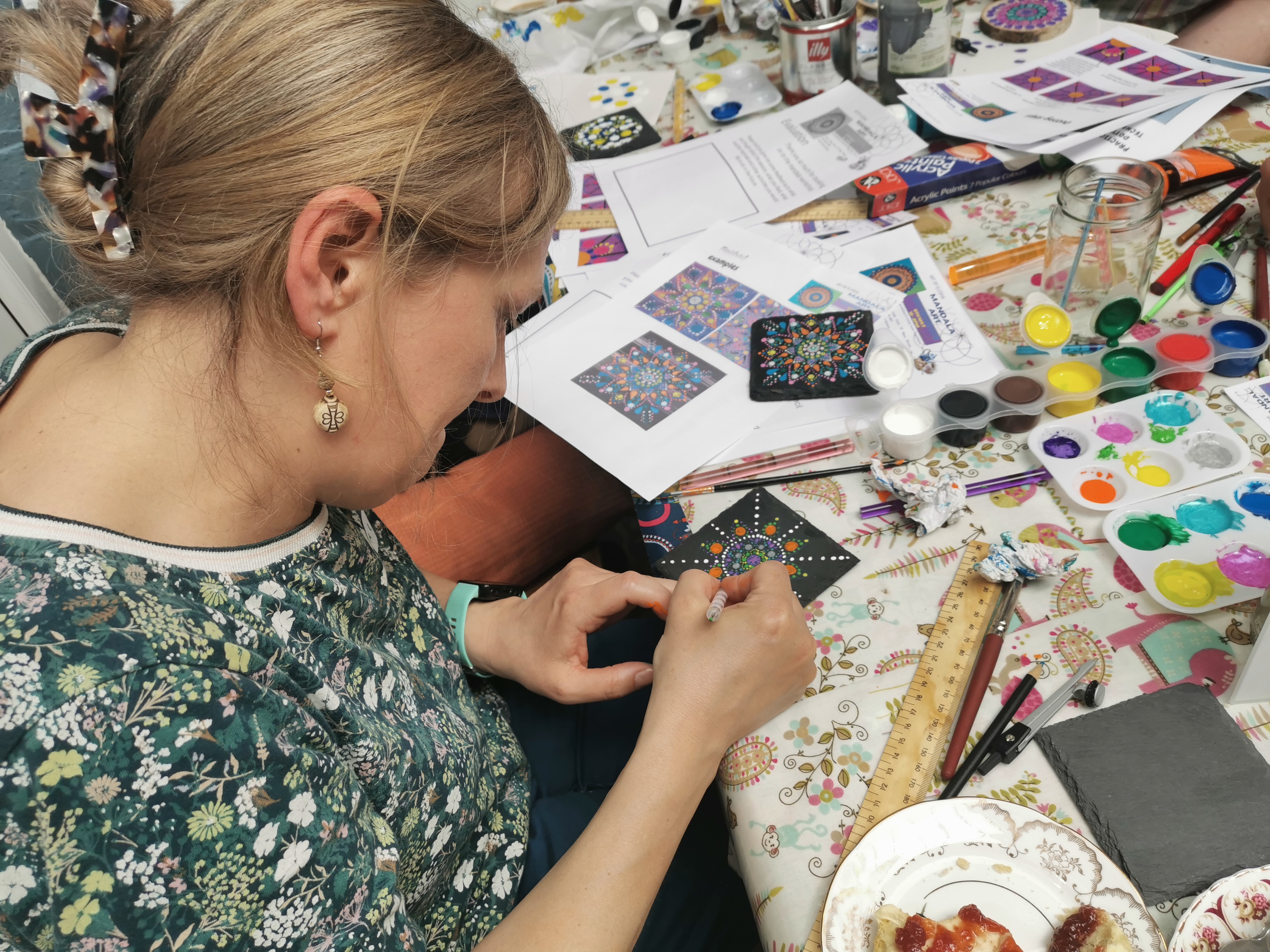 A person sits at a table covered with paints, brushes and printed mandala guides, carefully decorating a small black tile with colourful dotted patterns. Various paint pots, tools, finished example tiles and craft materials are spread across the patterned tablecloth.