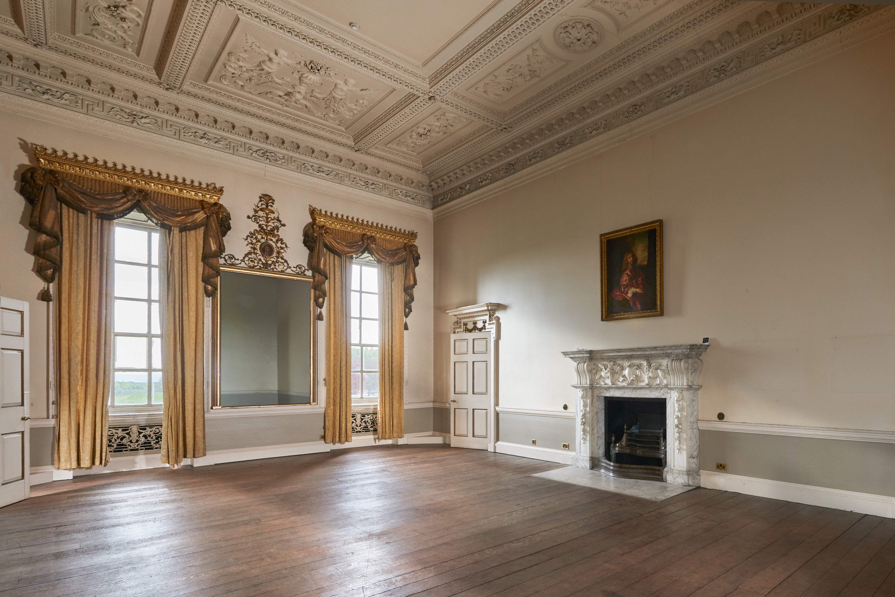 An elegant historic room with ornate plasterwork on the ceiling, tall windows draped with gold curtains, and a large decorative mirror between them. A marble fireplace with carved detailing sits on the right wall beneath a framed portrait. The room has plain painted walls and a wide wooden floor.