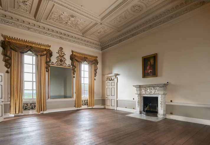 An elegant historic room with ornate plasterwork on the ceiling, tall windows draped with gold curtains, and a large decorative mirror between them. A marble fireplace with carved detailing sits on the right wall beneath a framed portrait. The room has plain painted walls and a wide wooden floor.