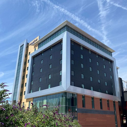 Modern multi-story building with a distinctive square frame design around its dark central section, featuring rows of windows. The building has beige and red exterior elements, with glass panels at the lower level. Clear blue sky with streaks of white clouds in the background and greenery in the foreground.