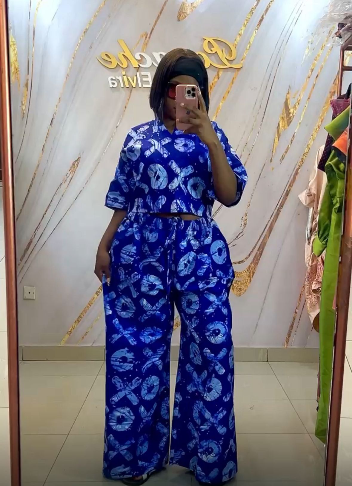A person wearing a vibrant blue two-piece outfit featuring a loose-fitting top and wide-leg trousers with bold white tie-dye patterns. The outfit is styled in a modern Afrocentric design and displayed in a boutique setting with a marble-effect wall and gold accents in the background.