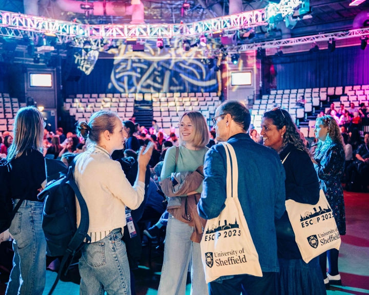 Large  area for conference. Dozens of attendees are gathered in groups, talking and holding event bags. Seating in the back ground.