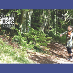 A woman stands in a forest, on a sunny day, playing violin.
