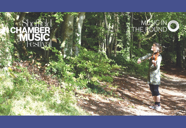A woman stands in a forest, on a sunny day, playing violin.