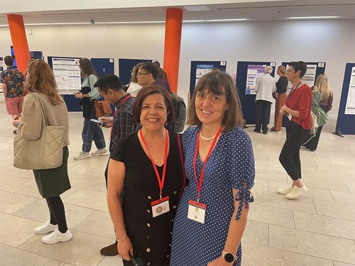 Two individuals wearing conference badges and red lanyards stand in the foreground of an indoor event space. Behind them, several people are engaged in conversation and viewing research posters displayed on dark blue boards. The setting appears to be a professional or academic conference with bright lighting and orange structural columns.