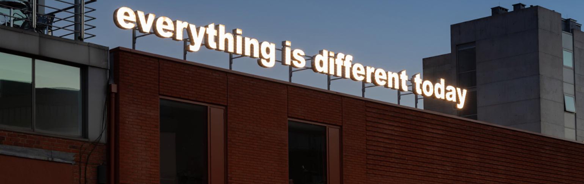 A red brick building. On the side is a sign that reads 'Site Gallery'. On the roof is an illuminated sign, made up of individual letters, that reads 'everything is different today'.