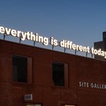A red brick building. On the side is a sign that reads 'Site Gallery'. On the roof is an illuminated sign, made up of individual letters, that reads 'everything is different today'.