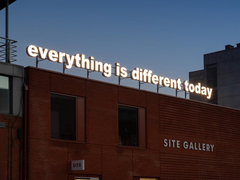 A red brick building. On the side is a sign that reads 'Site Gallery'. On the roof is an illuminated sign, made up of individual letters, that reads 'everything is different today'.