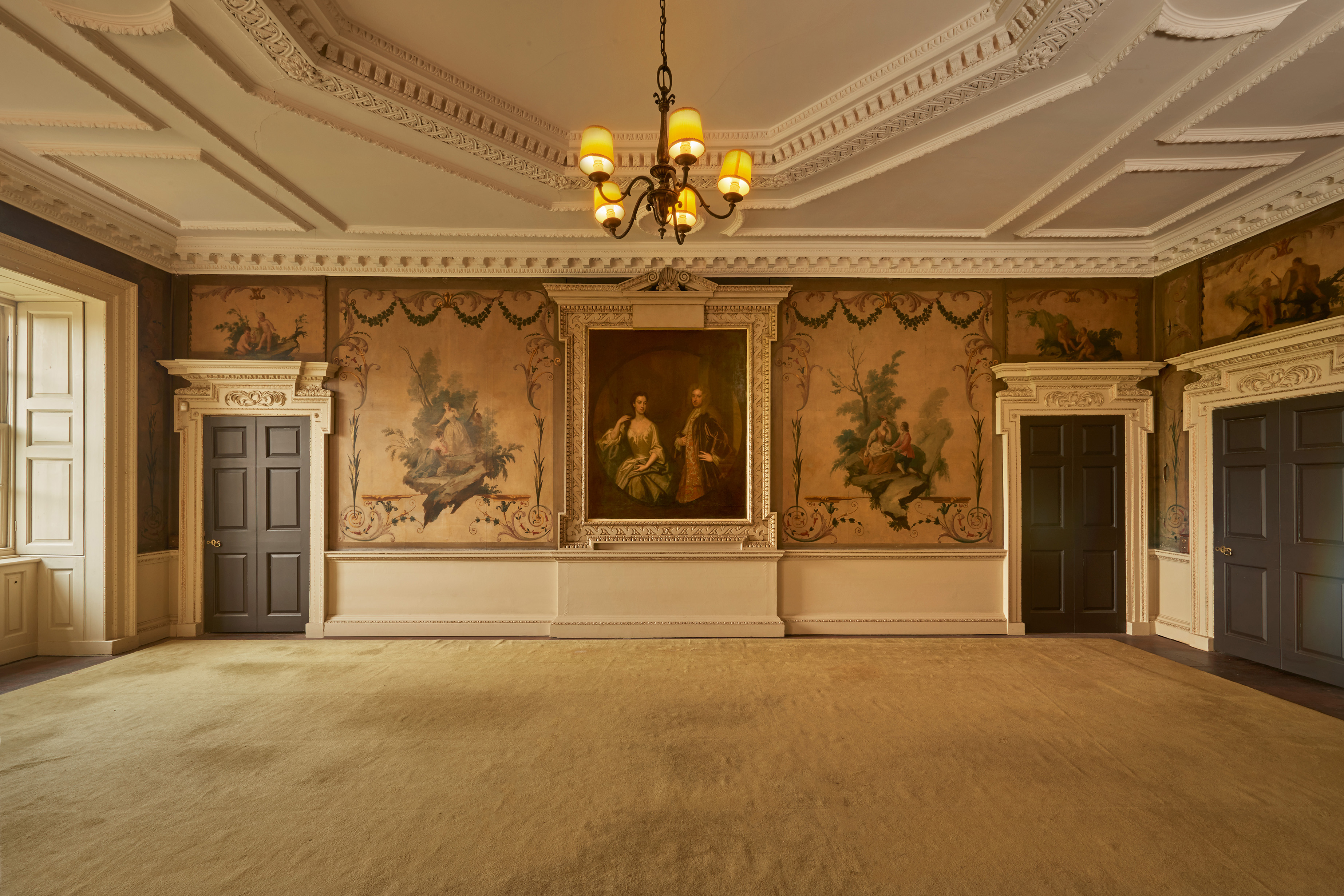 An ornate historic room with pale walls, decorative plasterwork on the ceiling, and a central chandelier. Large painted murals and a framed portrait adorn the walls, set between carved doorframes and dark wooden doors. Tall windows on the left let in soft natural light, illuminating the wide, empty carpeted floor.