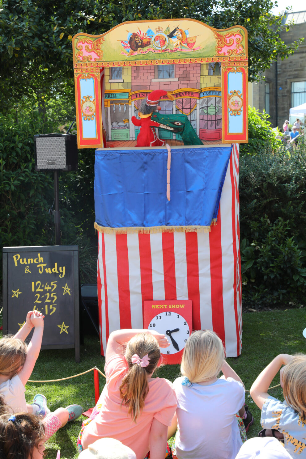 A traditional Punch and Judy puppet show takes place outdoors, with a striped red and white booth and colourful illustrated top panel. Two puppets, Punch in red and a crocodile, are visible inside the booth. A blackboard to the side lists show times, and a clock sign indicates the next performance. Several children sit on the grass watching, with greenery and part of a building in the background.