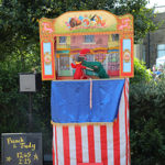 A traditional Punch and Judy puppet show takes place outdoors, with a striped red and white booth and colourful illustrated top panel. Two puppets, Punch in red and a crocodile, are visible inside the booth. A blackboard to the side lists show times, and a clock sign indicates the next performance. Several children sit on the grass watching, with greenery and part of a building in the background.