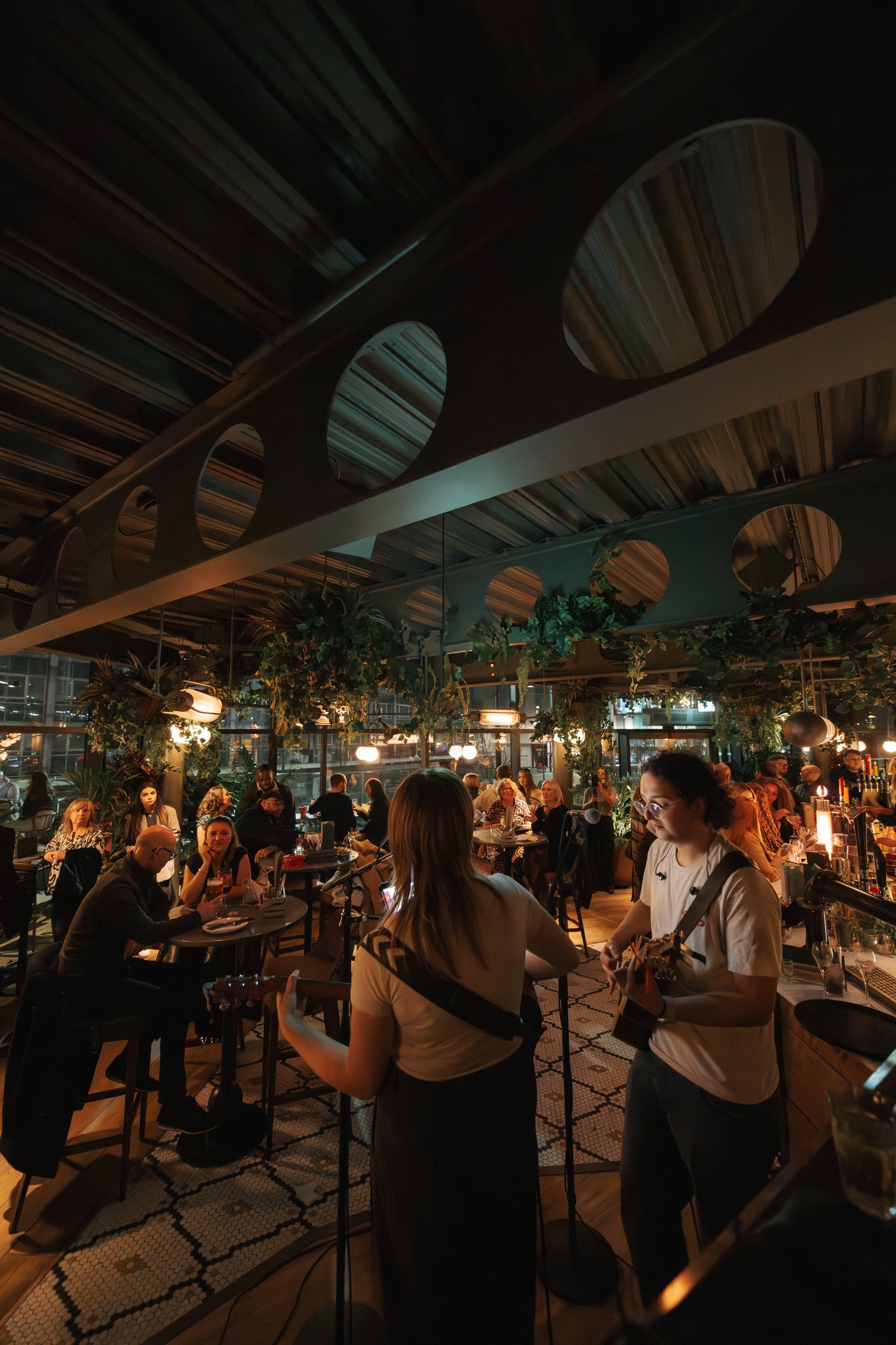 A lively indoor restaurant scene with musicians performing on a small stage and guests seated at tables, surrounded by hanging plants and warm atmospheric lighting.