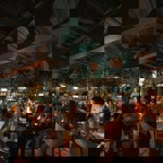 A lively indoor restaurant scene with musicians performing on a small stage and guests seated at tables, surrounded by hanging plants and warm atmospheric lighting.