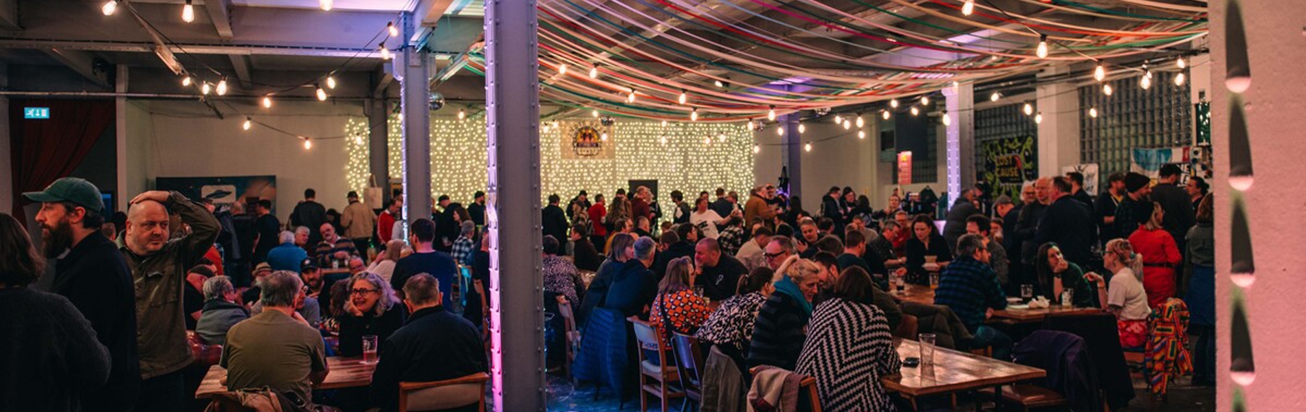A large indoor festival space filled with people seated at tables and standing in groups. Colourful ribbons hang across the ceiling, and string lights illuminate the room. The atmosphere appears busy and social, with many attendees enjoying drinks.