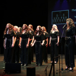 A vocal performance group stands on a dimly lit stage, arranged in a formation facing the audience. All performers wear matching black, sparkling outfits. A microphone on a stand is positioned to the right. Behind them, a large screen displays text and an image related to the performance. Credit: Green Vision Photography