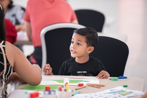 A child sitting at a table engaged in an arts and crafts activity. The table is covered with paper, colored pencils, glue sticks, and other craft supplies. Other people are seated at tables in the background in a bright indoor setting.