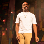 Person wearing a plain white short-sleeved T-shirt, beige trousers, and gold jewellery including a necklace and bracelet, standing on a stage. The background features a colourful, abstract mural with geometric shapes in warm tones and several small red lights. The scene is warmly lit, creating a theatrical atmosphere.