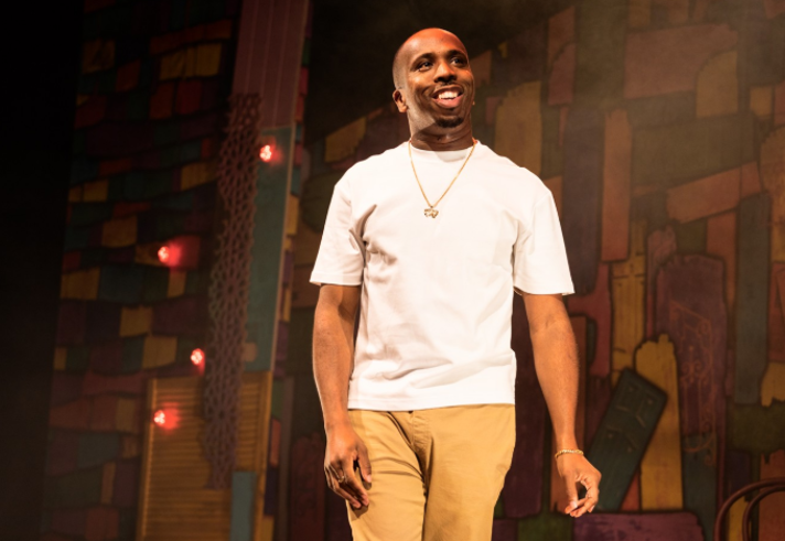 Person wearing a plain white short-sleeved T-shirt, beige trousers, and gold jewellery including a necklace and bracelet, standing on a stage. The background features a colourful, abstract mural with geometric shapes in warm tones and several small red lights. The scene is warmly lit, creating a theatrical atmosphere.