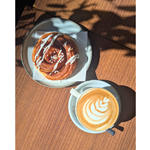 A wooden table, in the sunshine, with a danish pastry and a cappuccino.