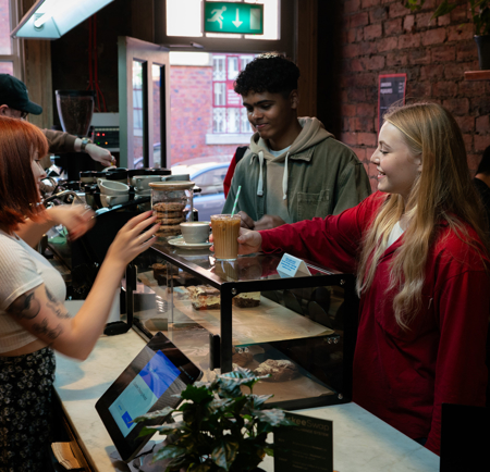 Barista serving a takeaway coffee to a customer inside a cozy café with exposed brick walls and wooden shelves displaying coffee bags. The counter features a glass display case with pastries, a small potted plant, and a payment terminal. A large window in the background lets in natural light, revealing a view of a red brick building outside.