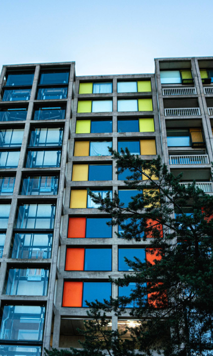 A brutalist high-rise apartment block made of concrete and glass. The windows are interspersed with bright panels in yellows and oranges.