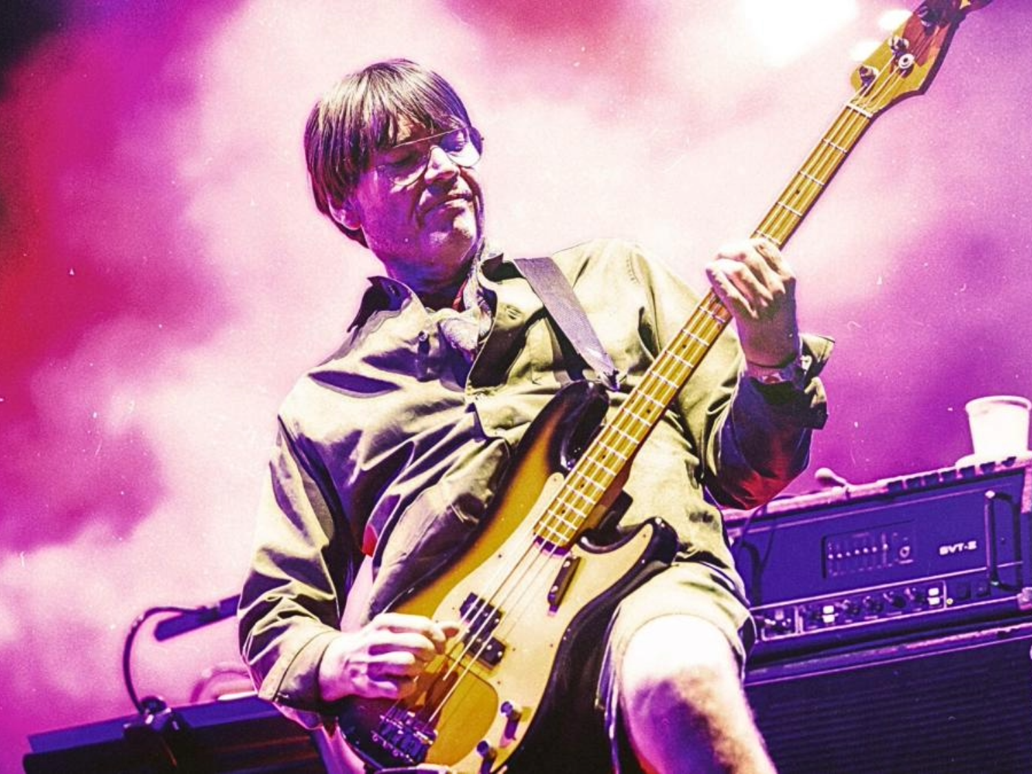A musician is performing on stage, playing an electric bass guitar. The person is wearing a long-sleeved shirt and shorts, with the guitar strap across the shoulder. The background is filled with vibrant pink and purple stage lighting, and there is audio equipment and a cup placed on top of an amplifier next to the performer.