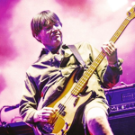 A musician is performing on stage, playing an electric bass guitar. The person is wearing a long-sleeved shirt and shorts, with the guitar strap across the shoulder. The background is filled with vibrant pink and purple stage lighting, and there is audio equipment and a cup placed on top of an amplifier next to the performer.