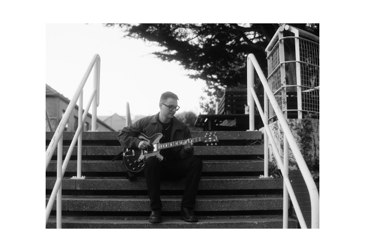 A person is seated on outdoor concrete steps, playing an electric guitar. The guitar has a semi-hollow body with a dark finish and visible f-holes. The individual is wearing a dark jacket and trousers, with both hands positioned on the guitar neck and body. White railings frame the steps on either side, and a tree with dense foliage is visible in the background along with a bench and part of a stone wall. The image is in black and white, creating a classic, moody atmosphere.