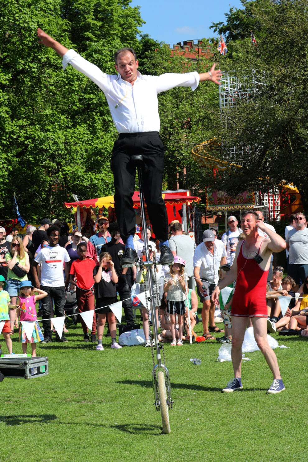 A performer balances on a very tall unicycle with arms outstretched during an outdoor event. Another performer in a red athletic outfit stands nearby, appearing to assist. Behind them, a large crowd watches, with colourful bunting, trees, and fairground stalls visible in the background on a sunny day.
