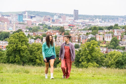 Two people walking on a grassy hill with a cityscape in the background, featuring rows of houses, trees, and tall buildings under an overcast sky.