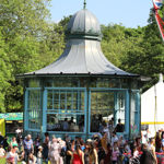 A decorative Victorian-style bandstand painted in blue stands in a park during a sunny outdoor event. People gather around and inside the structure, with groups chatting and walking on the grass. The background features lush green trees, a white marquee tent, and a tall flagpole with a Union Jack flag. The scene is lively, with festival activities taking place in a bright, open setting.
