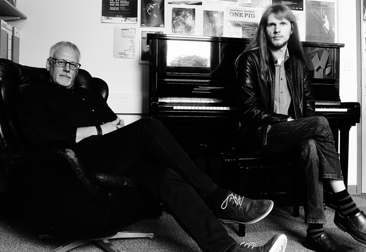 A black and white image of two people sat next to a piano.
