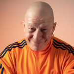 A picture of the performer wearing an orange tracksuit, with a light orange background. The performer is smiling and looking directly into the camera