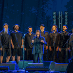 Fifteen men, standing on a stage in a semicircle, performing songs.