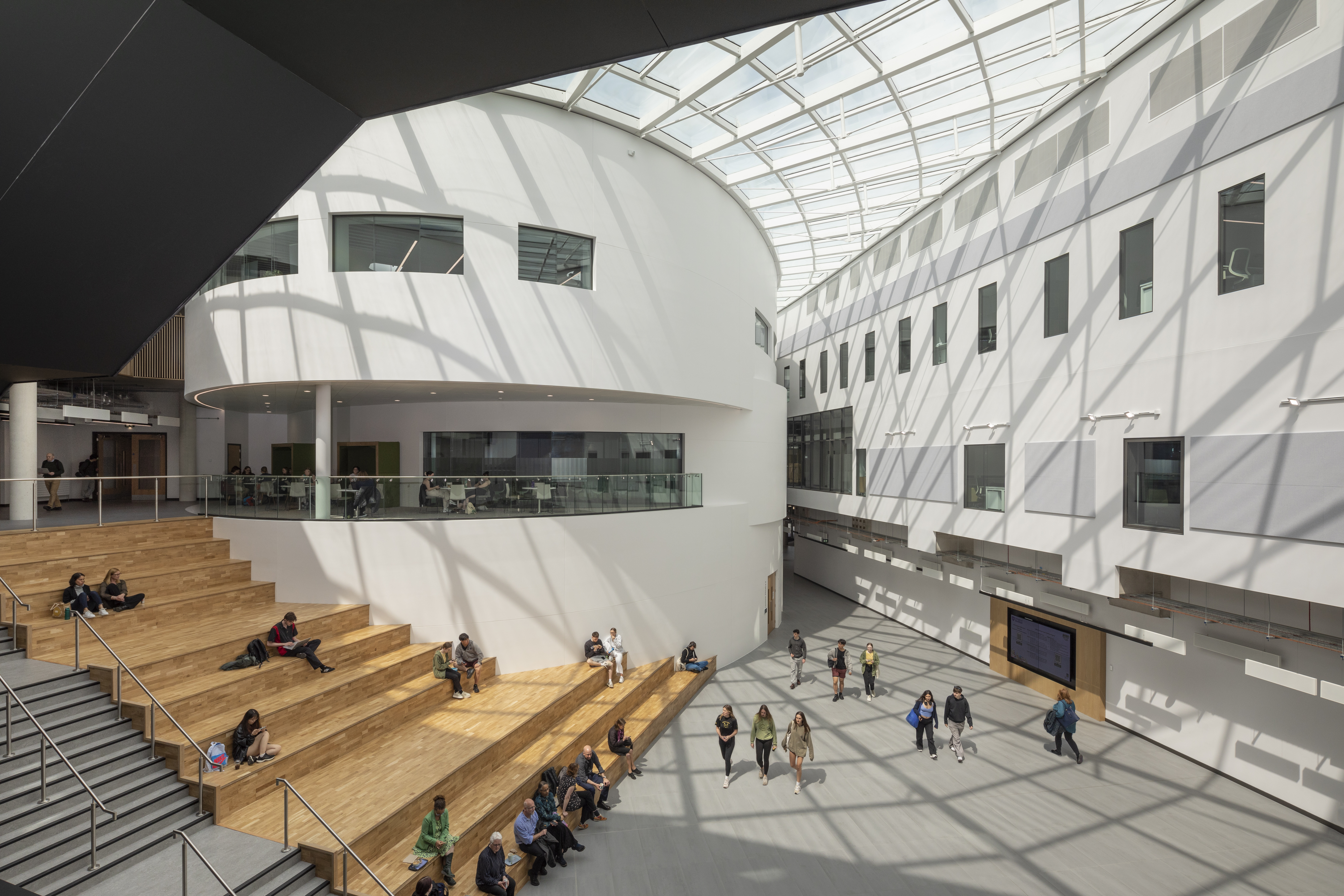 Spacious modern atrium with a curved white wall and glass ceiling allowing natural light to stream in. Wooden tiered seating occupies one side, where people are sitting and talking, while others walk across the open floor. The design features clean lines, large windows, and a bright, airy atmosphere.