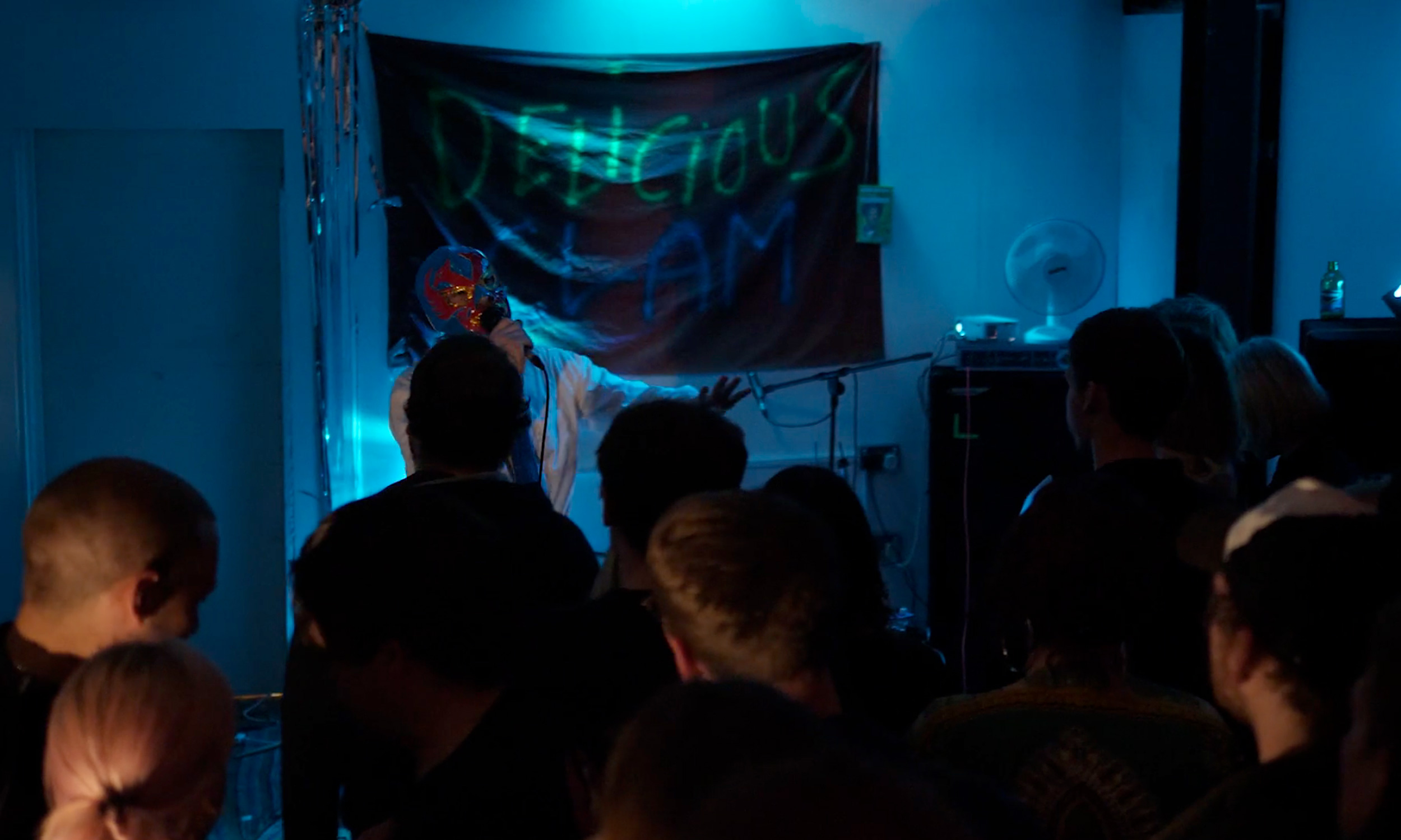 A performer wearing a bright, colorful mask and white outfit stands in front of a banner with neon-style text that reads “DELICIOUS CLAM.” The person is holding a microphone and gesturing toward the audience in a dimly lit room illuminated by blue lighting. Several people are gathered in the foreground, watching the performance, and metallic streamers hang along the wall to the left.
