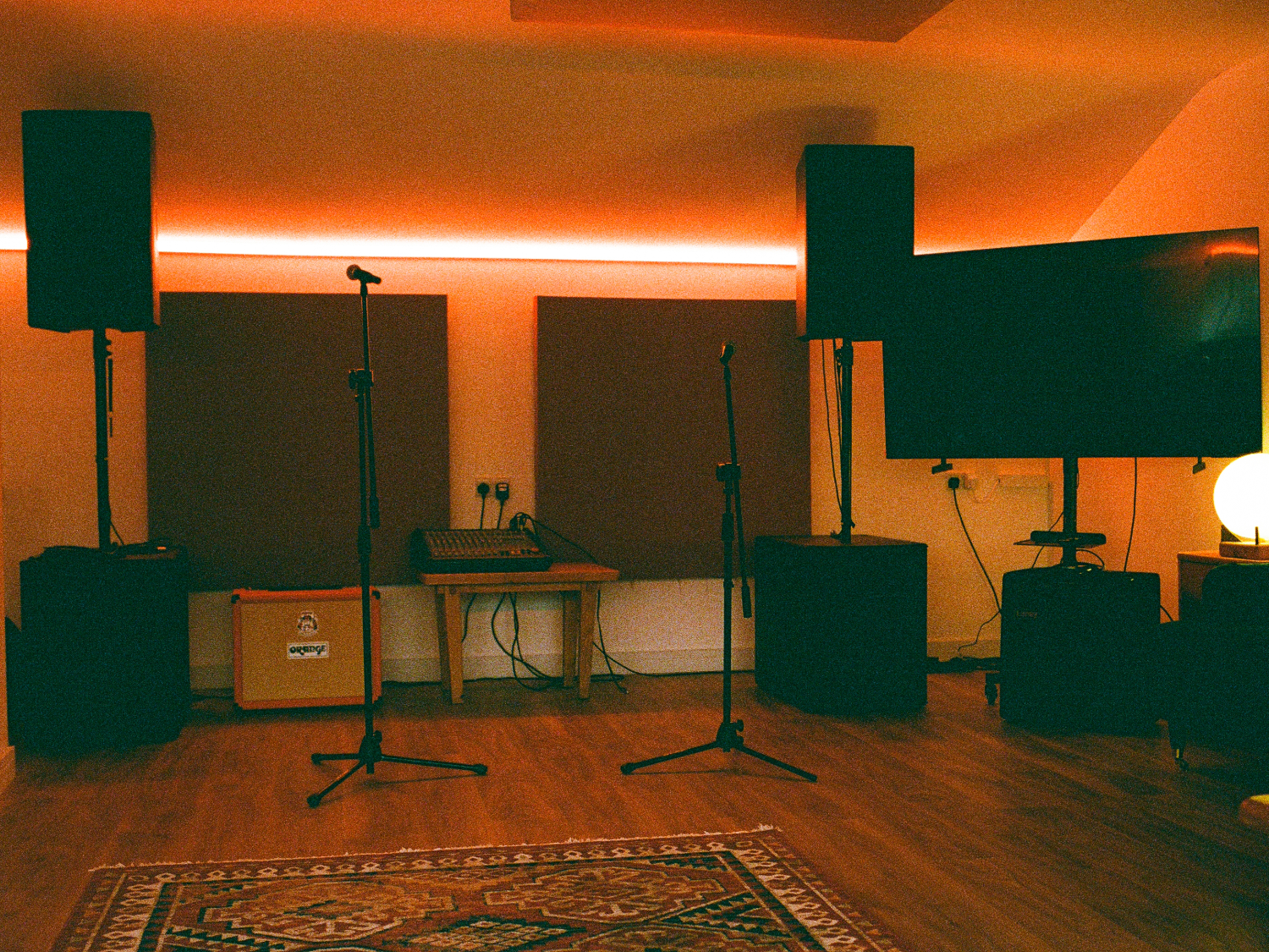 Music studio with warm ambient lighting, featuring two large speakers on stands, two microphone stands, and a flat-screen TV on the right. Acoustic panels are mounted on the back wall above a small wooden table holding audio equipment. An orange guitar amplifier sits on the floor to the left. The room has wooden flooring and a patterned rug in the foreground, creating a cosy, professional recording space.