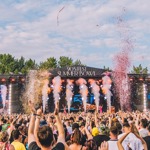 A lively festival crowd faces the “90’s Fest Summer Bowl” stage as smoke cannons shoot white plumes and colorful confetti fills the air. People have their hands raised, and the scene is bright and energetic with trees and blue sky in the background.