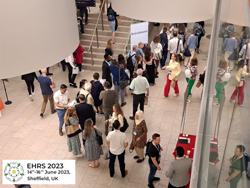 An indoor conference venue with a large group of people gathered in an open area near a staircase. Attendees are standing, walking, and conversing around exhibition tables and banners, including one with the text “Reichenschwand.” The space features modern architecture with curved white walls, glass panels, and bright lighting. A sign in the bottom-left corner reads “EHRS 2023, 14th–16th June 2023, Sheffield, UK.”