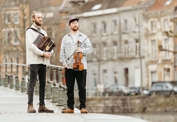 Hartwin Dhoore & Ross Grant hold their instruments, standing on a bridge in an urban setting.