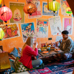 Two people are seated on cushions on the floor, at a low table, enjoying the meal. There are brightly coloured lanterns hanging from the ceiling. The orange walls are decorated with old and faded posters.