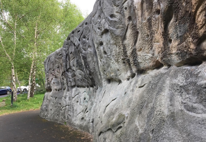 Outdoor climbing wall with various handholds and textures. It is located beside a paved path, surrounded by grass and trees, with parked cars visible in the background.