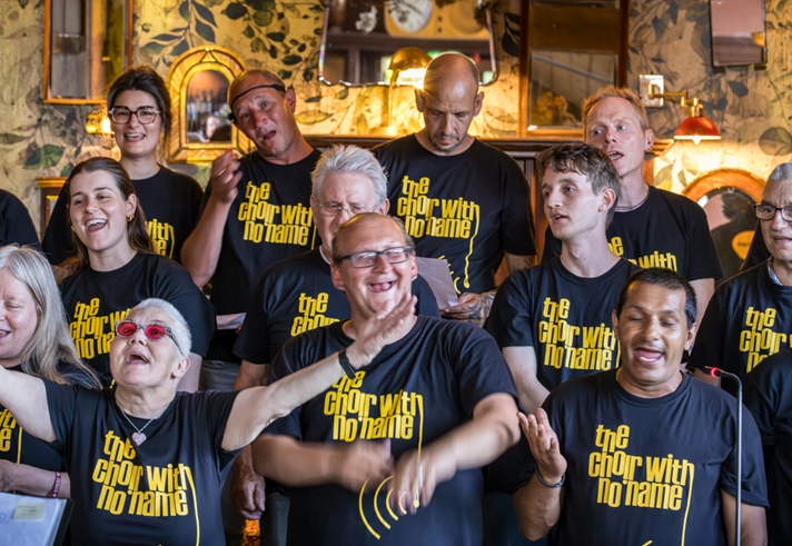 Members of The Choir With No Name singing together in black t-shirts with yellow text, standing in front of a decorative wall with mirrors and lights.
