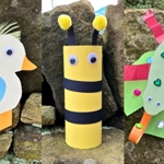 A photo collage showing three colourful children’s craft animals made from paper and recycled materials, displayed outdoors against stone rocks. On the left is a white bird with a yellow beak, red feathered wing, blue circle body detail, and orange legs. In the centre is a yellow and black striped bee made from a cardboard tube, with two googly eyes and two yellow pom‑pom antennae. On the right is a green bug made from layered paper shapes with red stripes, decorated with sequins and a long green paper head