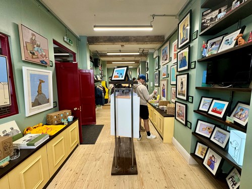 Interior of an art gallery or shop with light wooden floors and green walls covered in framed illustrations and prints. Shelves display artwork, books, and decorative items. A central stand holds a tablet, and the space leads to a red doorway at the back.