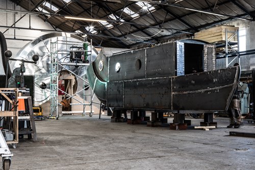 Interior of a large industrial workshop with high ceilings and metal beams, showing a partially constructed steel structure resembling a boat hull on supports. In the background, scaffolding surrounds a large circular metallic component, with tools and equipment scattered around the concrete floor.