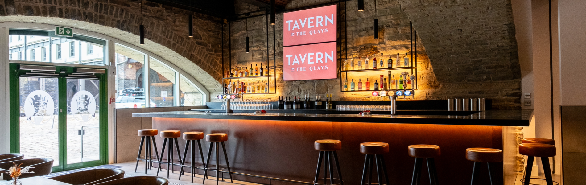 The bar at Tavern on The Quays, with the vaulted stone ceiling plus bar stools lined up along the bar and tables and chairs in the foreground.