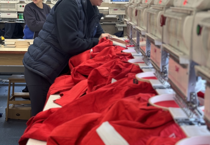 Person wearing ear protection and a padded jacket operating a row of embroidery machines in a workshop. Bright red garments are laid out on the machines for stitching. Another person is visible in the background, and the workspace includes tables and equipment under bright indoor lighting.