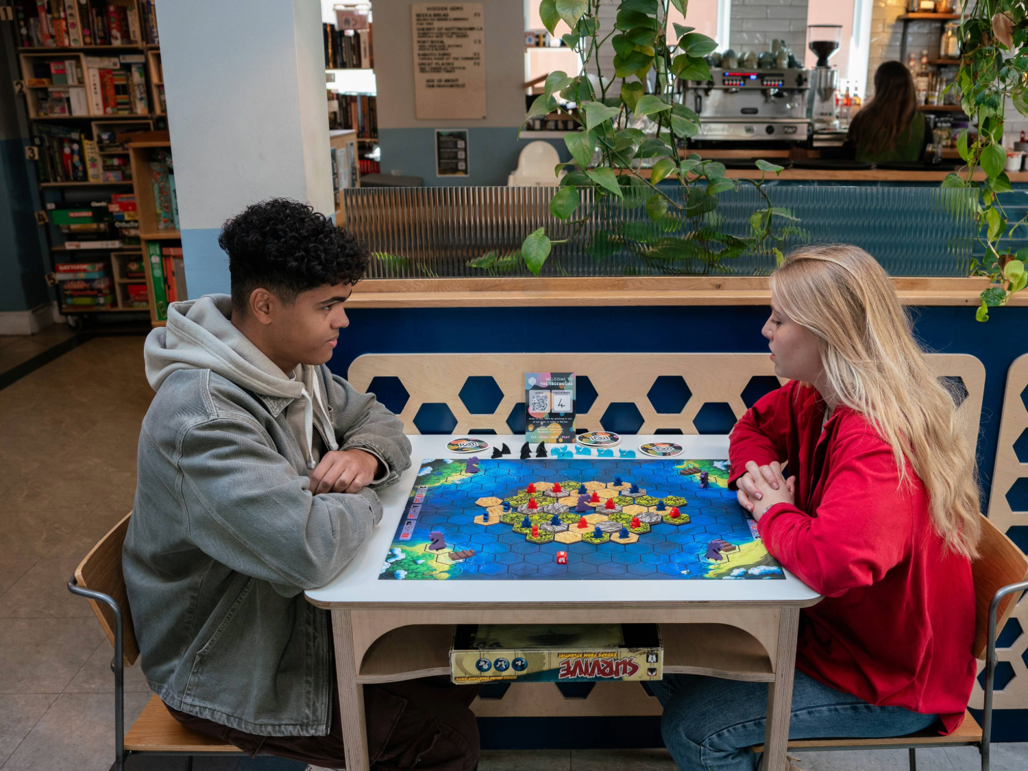 Two people are sat at a table, in a cafe, playing a board game and drinking coffees. Behind them is shelving stacked full of all different board games, lots of leafy plants and other tables filled with people.