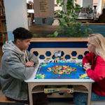 Two people are sat at a table, in a cafe, playing a board game and drinking coffees. Behind them is shelving stacked full of all different board games, lots of leafy plants and other tables filled with people.